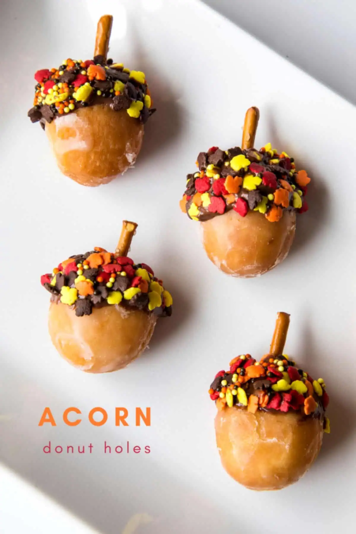 donut holes dipped about 30% in chocolate with fall sprinkles in the chocolate and a pretzel stick in the top to make donut hole acorns