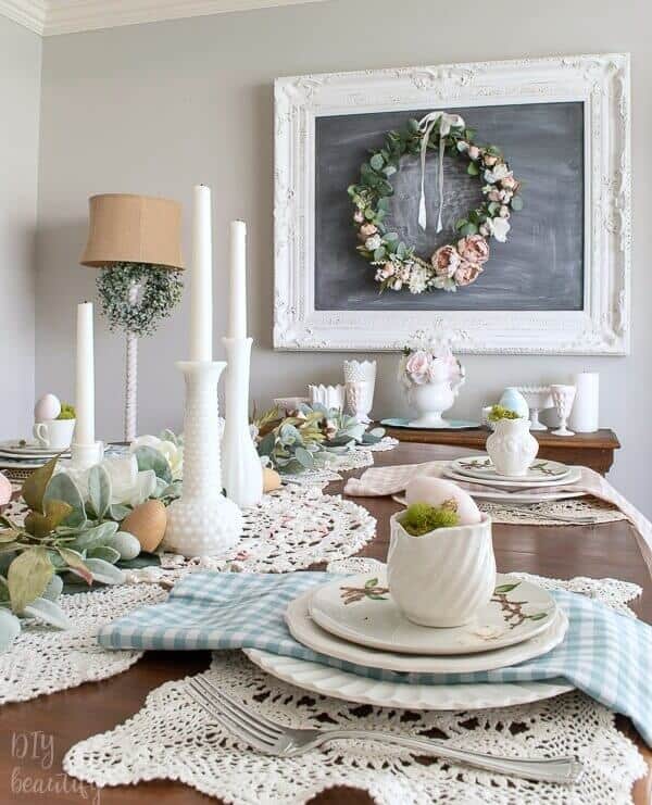 Easter Tablescape ideas, doiily table runner with milk glass vases, candles, greenery and eggs, each place setting layered with white dishes and blue and pink gingham napkins.