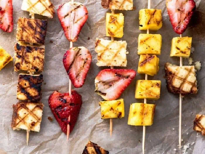 Skewers with fresh pineapple, strawberries and squares of cake being grilled for camping.