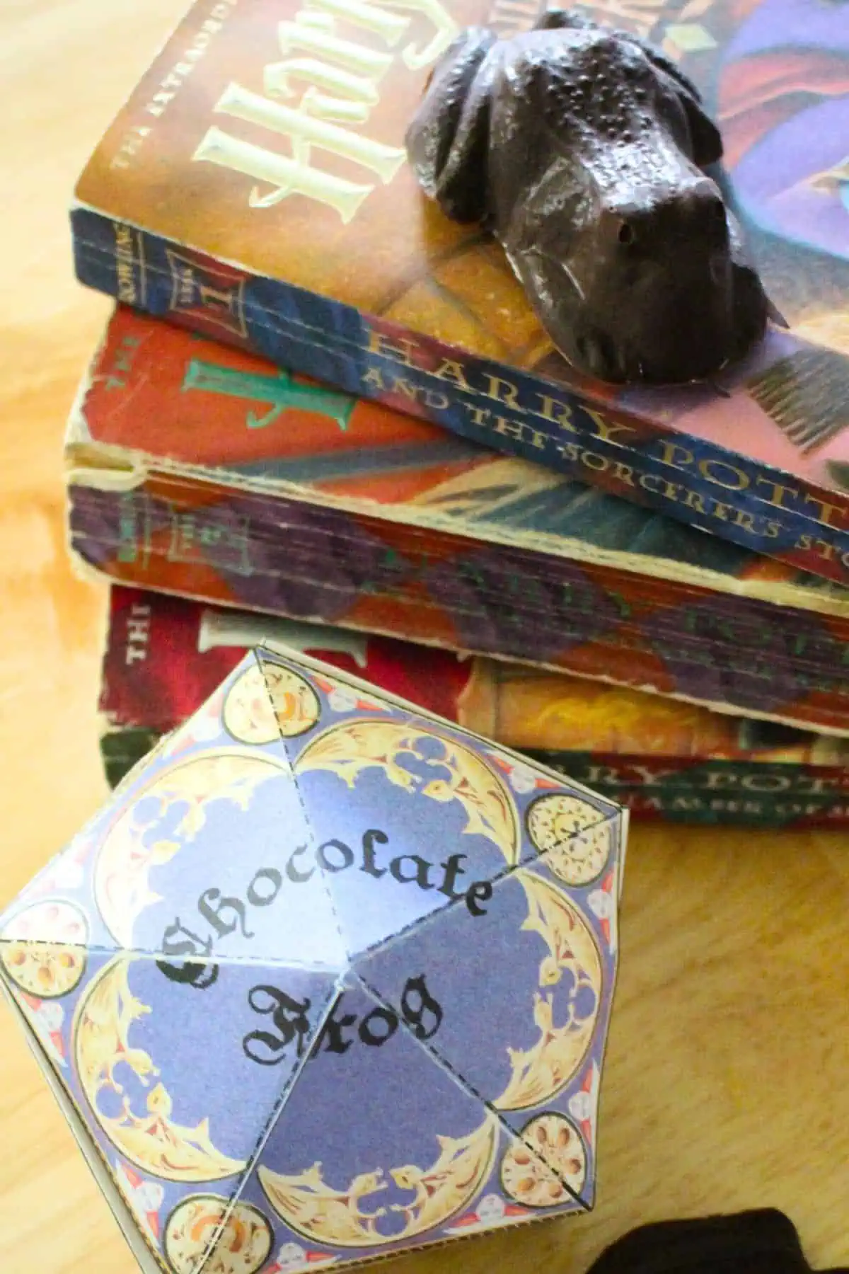 Harry potter books stacked with a chocolate frog on top, a chocolate frog box to the side