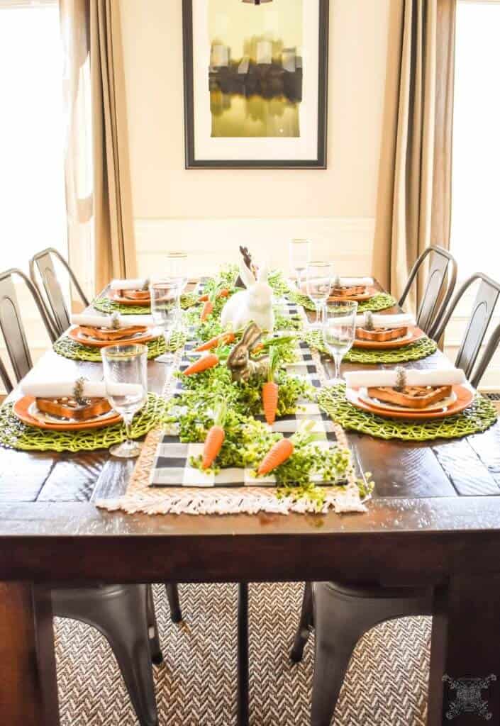 Green straw placemats at each place setting layered with orange and white plates. Down the center of the table are layered 2 tablerunners, one black and white buffalo check and natural woven runner. Carrots, greenery and bunnies are scattered down the center of this farmhouse table.
