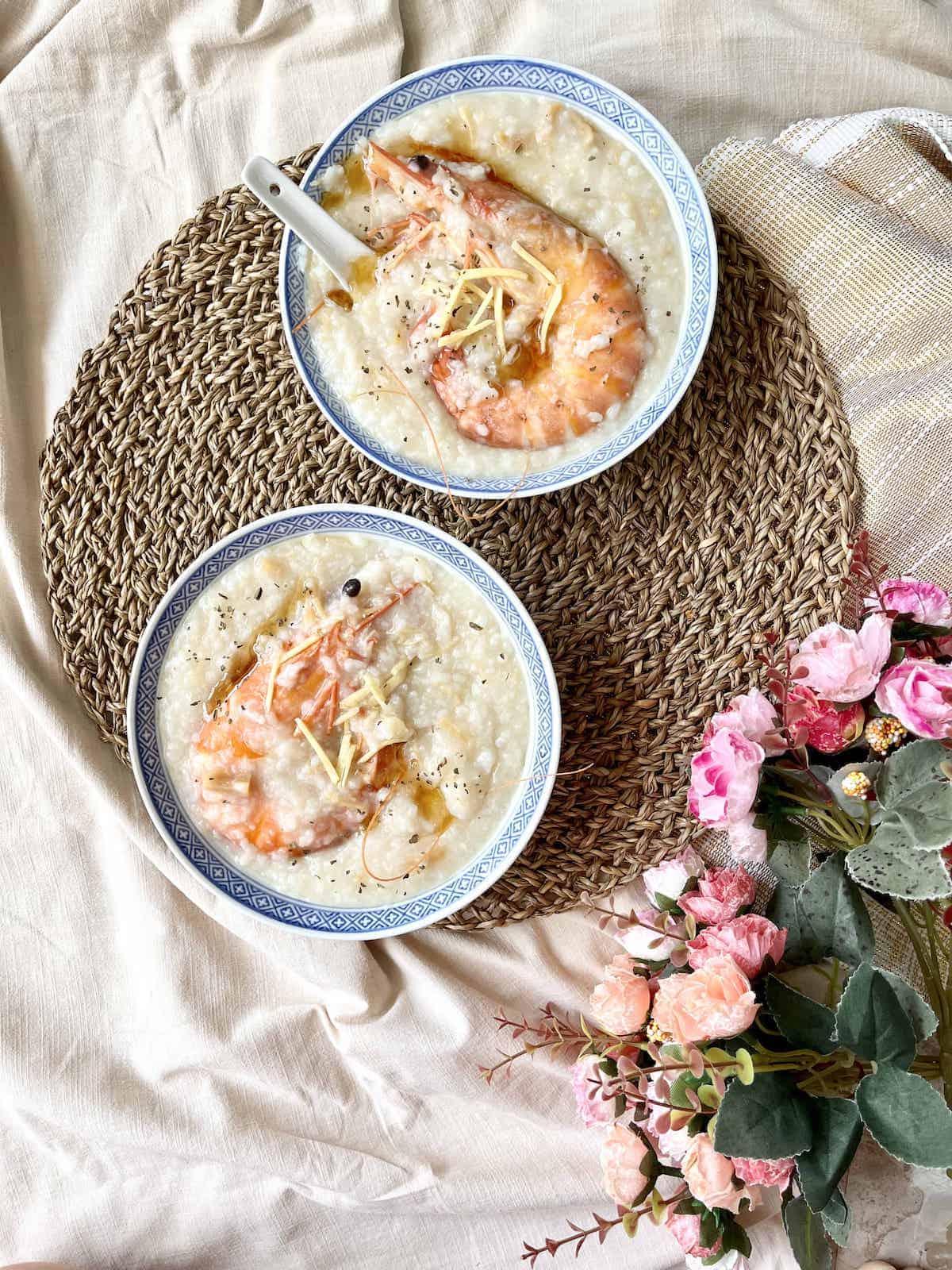 Two bowls of easy shrimp congee in a bowl with large prawn with flowers in the background.