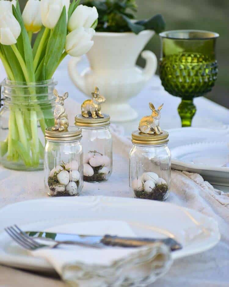 Easter Tablescape ideas, white tablecloth, white, plates and napkin, with vintage silverware, green stemware, mason jar filled with white tulips. Small mason jars filled with moss, easter eggs, topped with a gulded bunny.