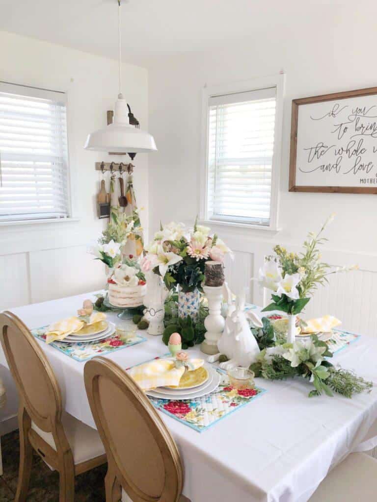 Easter Tablescape ideas, floral placemats, with white and yellow dishes, and white and yellow gingham napking, centerpiece of candles, bunnies, greenery and faux cake.