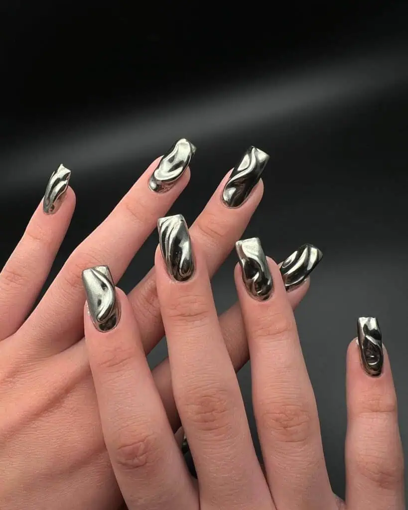 Hands displaying different metallic nail styles, featuring black nails with silver specks, dark gold polish, and shiny red acrylic nails.